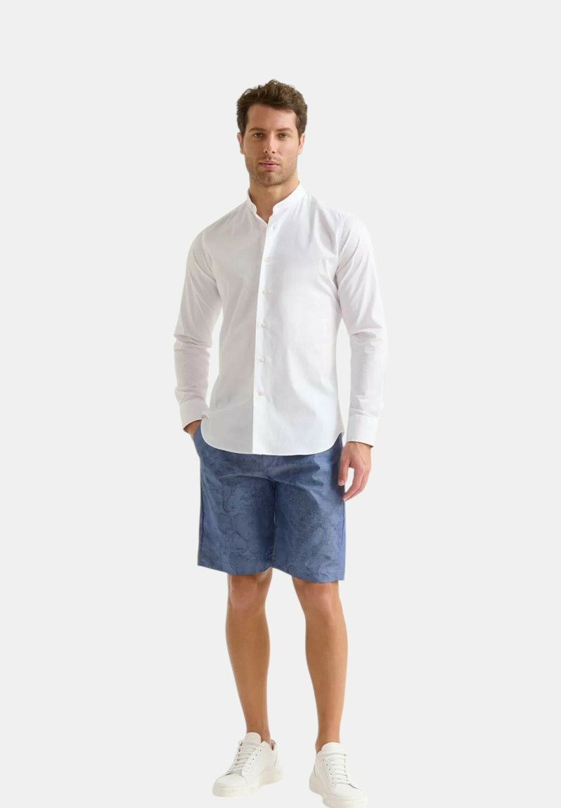 Camicia bianca a maniche lunghe con collo alla cinese, abbinata a pantaloni corti blu a fantasia e sneaker bianche, in piedi su uno sfondo neutro.