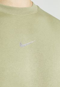Ljus olivgrön t-shirt med rund halsringning och liten silverfärgad Nike-swoosh-logotyp på bröstet, visad från halsen till övre torso.