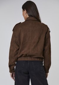 Brązowa zamszowa kurtka z kołnierzem, naramiennikami oraz elastycznym pasem. Posiada długie rękawy i luźny krój.