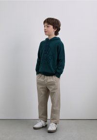 Grön huvtröja med en mörkblå dödskalle och korslagda ben-grafik, kombinerad med beige byxor och vita sneakers, stående på ett grått golv.