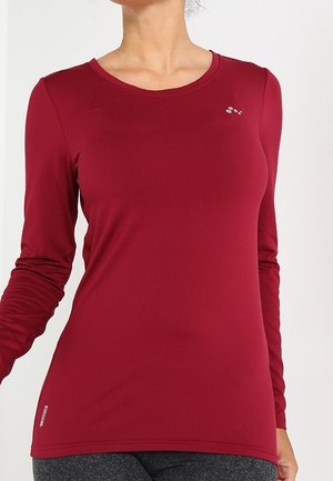 Žena má na sobě přiléhavé bordó sportovní tričko s dlouhým rukávem a tmavě šedé legíny, stojí před světlým pozadím.