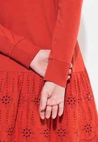 Rotes Langarmoberteil mit gerippten Bündchen, kombiniert mit einem Rock, der bestickte florale Muster und ausgeschnittene Details aufweist, alles in passenden Farbtönen.