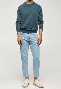 Türkisfarbener Pullover, hellblaue Jeans und weiße Turnschuhe mit beigefarbenen Sohlen. Der Pullover hat gerippte Bündchen und Saum, eine lockere Passform und ein schlichtes Design.