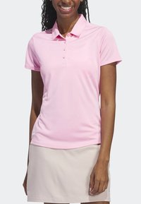 Femme souriante portant un polo à manches courtes rose clair et une jupe beige, sur fond uni.