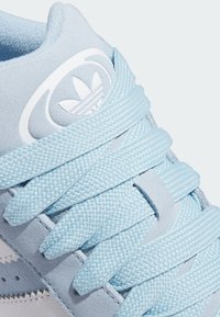 Light blue suede sneaker with textured surface, featuring flat braided laces and a white logo accent on the tongue and side.