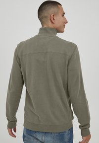 Felpa verde khaki con colletto alto, polsini e orlo a coste, e una texture liscia, caratterizzata da un design semplice e senza motivi visibili.