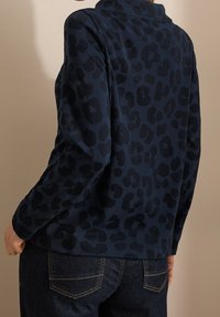 Maglietta a maniche lunghe blu navy con motivo maculato nero. Presenta un colletto e una vestibilità morbida, abbinata a jeans scuri. Tessuto morbido e testurizzato.