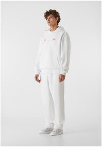 Hvit hettegenser med rosa logoer, har lomme foran og snor i hetten, kombinert med matchende hvite bukser og sneakers med grå detaljer.