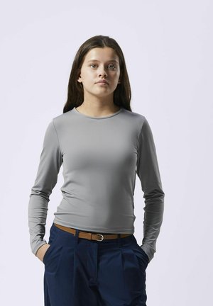 Langsleeve, grijs shirt van gladde stof met een ronde hals. Gecombineerd met blauwe broek en een bruine riem, handen in de zakken.