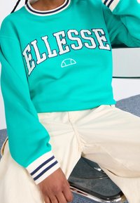 Turquoise trui met het "ELLESSE" logo in het wit, voorzien van een navy en witte gestreepte geribbelde kraag en manchetten, gedragen met lichtgekleurde broek.