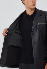 Zwarte leren jas met een afgekante kraag, voorzien van verticaal gestreepte voering, binnenzak en voorzijde met rits. G gedragen over een zwarte top.