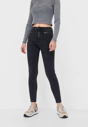 Jean skinny noir taille haute avec une texture lisse, poches avant et coupe ajustée, associés à un haut gris à manches longues cropped.