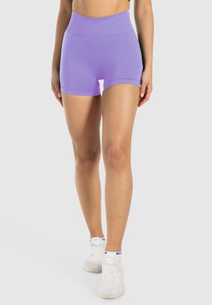 Vrouw in hoge naadloze lavendel shorts en witte sportschoenen, staand tegen een effen lichtgrijze achtergrond.