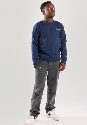 Dunkelblaues Crewneck-Sweatshirt mit kleinem weißen Logo, kombiniert mit grauen Hosen und weißen Sneakern. Einfaches Design, Baumwollmaterial.