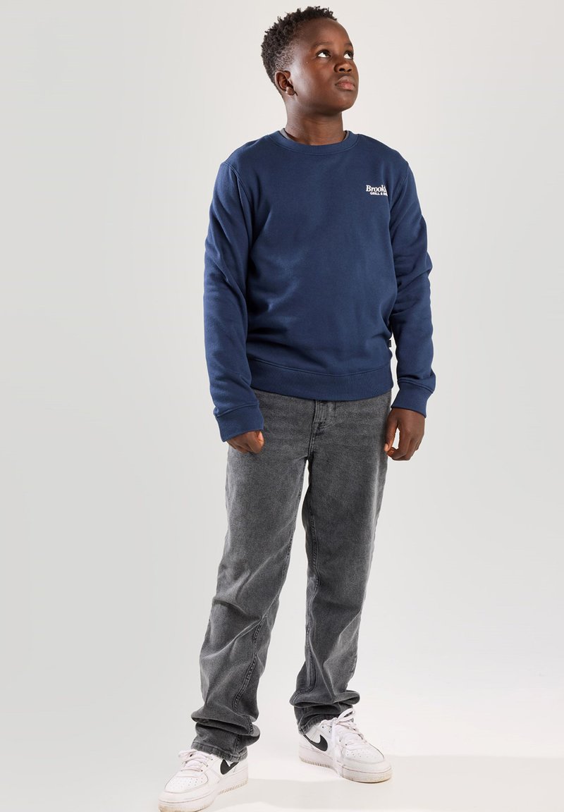 Sudadera de cuello redondo azul marino con un pequeño logo blanco, combinada con pantalones grises y zapatillas blancas. Diseño sencillo, material de algodón.