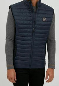 Navy puffer vest with horizontal quilted design, featuring a front zipper and logo patch. Lightweight fabric with a smooth texture.