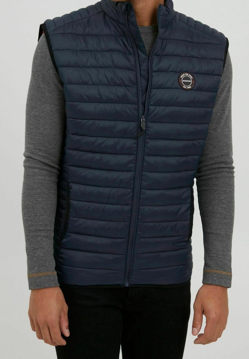 Navy puffer vest with horizontal quilted design, featuring a front zipper and logo patch. Lightweight fabric with a smooth texture.