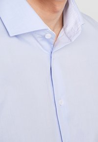 Jasnoniebieska koszula w jednolitym kolorze z prążkowanym, wewnętrznym kołnierzykiem. Ma miękką fakturę i subtelny połysk, zapinana na białe guziki.