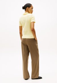Gulstribet polo-shirt med korte ærmer, parret med beige vide bukser. Modellen står med hænderne i lommerne, set bagfra.