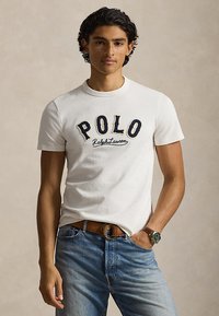 Νέος άνδρας που φοράει λευκό μπλουζάκι Polo Ralph Lauren, μπλε τζιν, καφέ ζώνη και ρολόι χειρός με πράσινο καντράν, στέκεται με το ένα χέρι στην τσέπη.