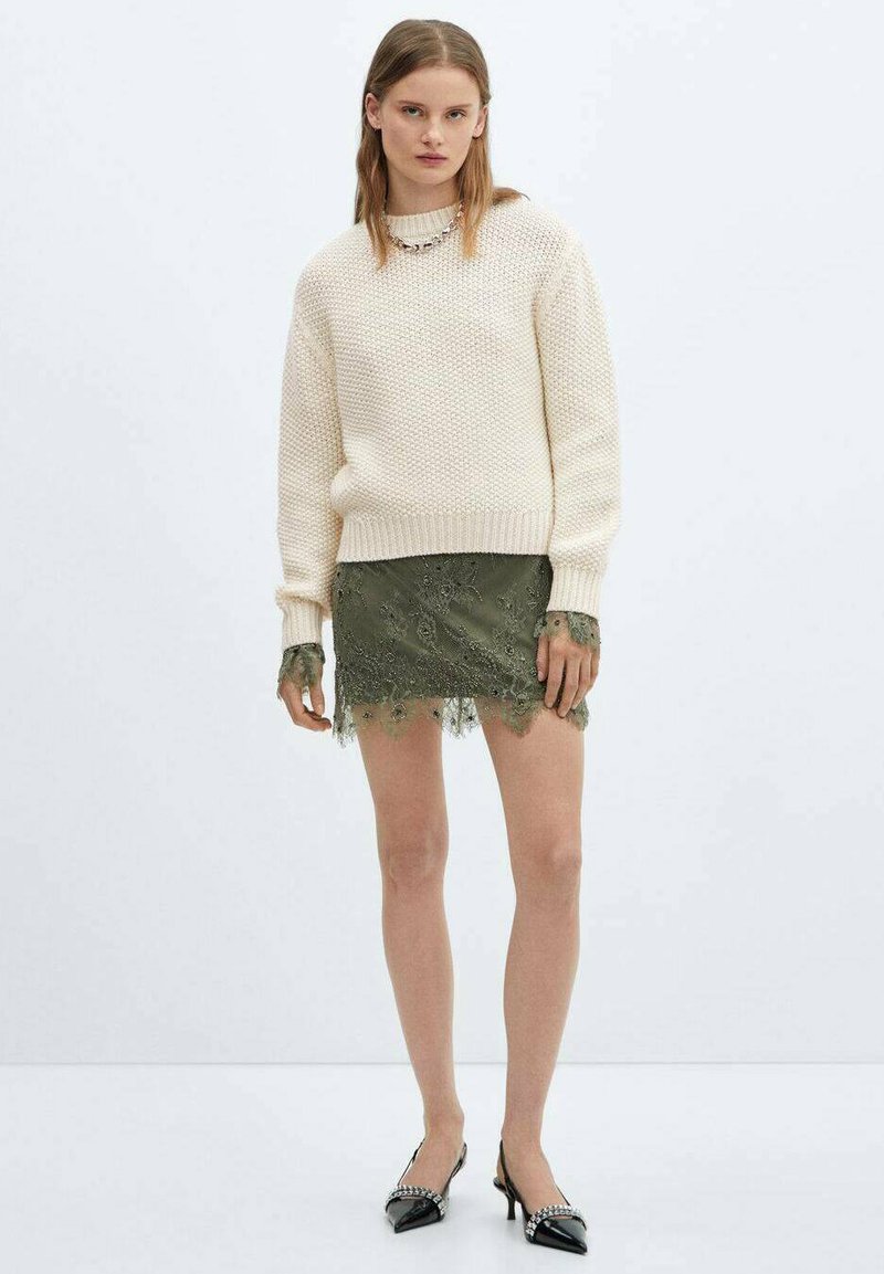 Pull en maille crème avec un motif texturé, associé à une jupe en dentelle vert olive ornée de broderies florales et à bord festonné. Talons noirs.