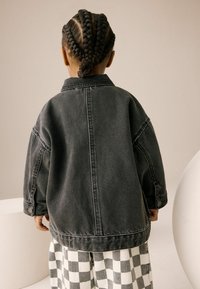 Veste en denim noire surdimensionnée avec un col et des poignets à boutons, portée par-dessus un pantalon à carreaux. Cheveux tressés visibles de dos.