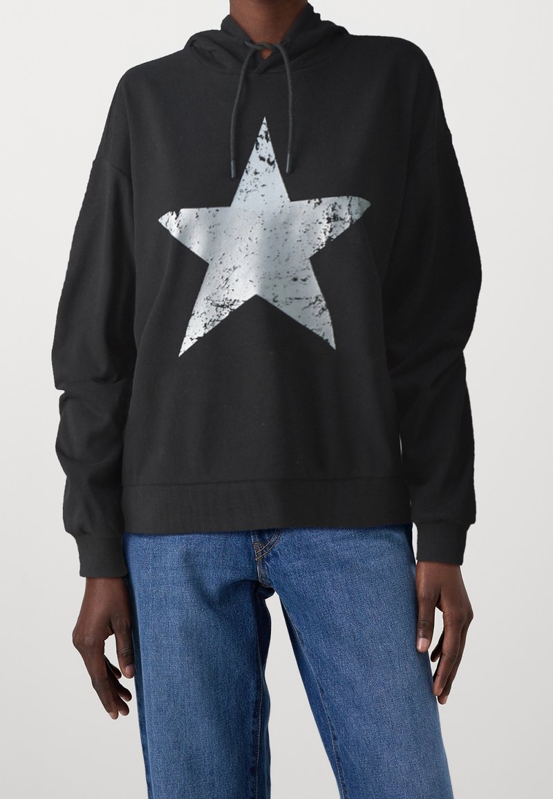 Personne portant un sweat à capuche noir avec une grande étoile argentée usée sur le devant, associée à un jean bleu, debout devant un fond uni.