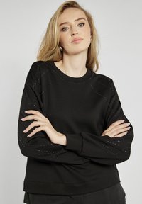 Czarna bluza z miękkiego materiału, z pikowanymi detalami na ramionach z wbudowanymi cyrkoniami. Okrągły dekolt i długie rękawy.