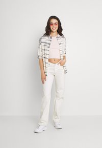 Modelo vestindo uma camisa com estampado tie-dye em branco e cinza, um crop top rosa claro e jeans brancos com corte evasê, além de sapatilhas brancas. Óculos de sol rosa claro.