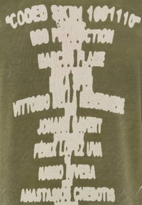 Izbalējis balts teksts ar neregulāru formu uz olīvu zaļas auduma, detaļas lielākoties nesalasāmas bojājuma un deformācijas dēļ.