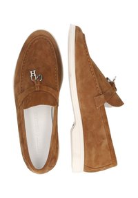 Scarpe senza lacci in camoscio marrone con suola in gomma bianca, caratterizzate da un piccolo fiocco e un accento metallico con la scritta "H2". Texture liscia, design classico.