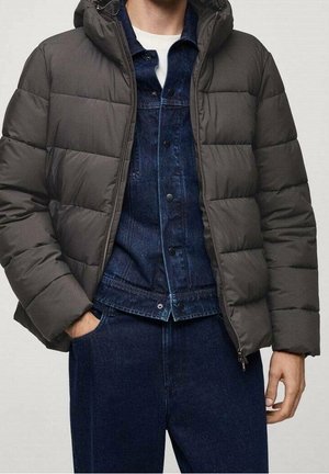 Homme portant une doudoune gris foncé par-dessus une veste en jean bleue et un pantalon en jean bleu, avec une main dans la poche et l'autre détendue.