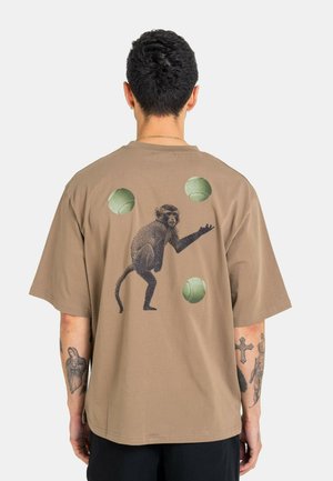 Person wearing a brown T-shirt with a graphic of a monkey juggling three green balls on the back, visible tattoos on both forearms.