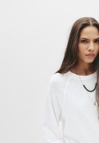 White long-sleeve shirt with a relaxed fit, featuring a subtle texture. Worn with layered necklaces, one with dark beads and another pendant.