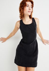 Μαύρο τζην φόρεμα με εφαρμοστή σχεδίαση, στρογγυλή λαιμόκοψη και ορατές ραφές. Το ύφασμα έχει ελαφρώς υφή και είναι μέχρι το γόνατο.