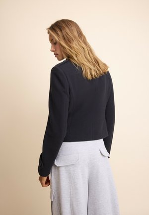 Femme aux cheveux blonds mi-longs portant une veste noire cintrée et un pantalon gris clair, debout devant un fond beige uni, vue de dos.