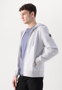 Sweatshirt de zíper cinza claro com um bolso frontal e punhos canelados. Usado sobre uma t-shirt azul clara, apresentando um logotipo circular na manga.