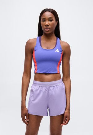 Femme aux longs cheveux foncés portant un crop top de sport violet avec des rayures rouges et un short violet clair, debout devant un fond uni.