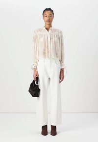 Blusa de gasa beige con estampado floral, con cuello y puños con volantes, combinada con pantalones anchos de cintura alta blancos y botas de tobillo marrones, sosteniendo un bolso negro.