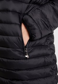 Chaqueta de plumas negra con secciones acolchadas, que presenta un bolsillo con cremallera y tirador de cremallera redondo. Tejido suave y brillante con una textura cálida.