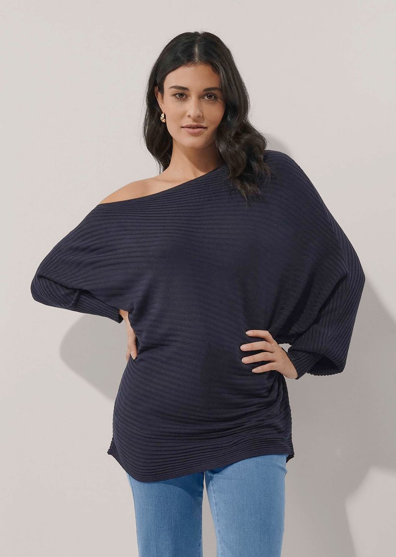 Frau mit dunklem welligem Haar trägt einen schulterfreien, gerippten marineblauen Pullover und hellblaue Jeans, steht mit den Händen in der Hüfte vor einer schlichten Wand.