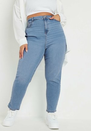 Jeans de tiro alto azul con un diseño ajustado y bolsillos frontales, que cuentan con un cierre clásico de botón y cremallera. Llevados con zapatillas blancas.