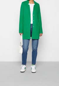 Abrigo de mezcla de lana verde con cuello, cierre de un solo botón, combinado con una camiseta blanca, jeans azules y zapatillas blancas con rayas verdes.