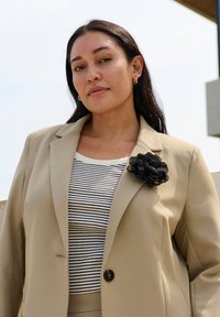 Blazer beige ajusté porté sur un haut rayé noir et blanc, avec un épingle de fleur noire texturée. Tissu lisse avec une forme structurée.