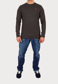 Gray long-sleeve shirt with round neck and small red logo, paired with blue denim jeans and black sneakers.