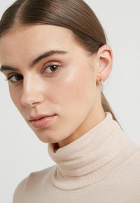 Gold knot stud earrings worn by a model with straight brown hair, featuring a knit turtleneck top in a light beige color.