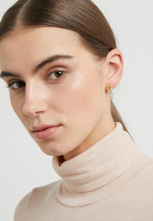 Gold knot stud earrings worn by a model with straight brown hair, featuring a knit turtleneck top in a light beige color.
