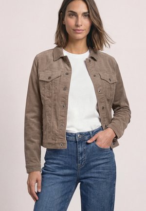 Femme portant une veste taupe boutonnée sur une chemise blanche et un jean bleu, une main dans la poche, sur un fond uni.