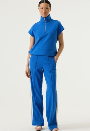 Blauw sportief setje bestaande uit een top met korte mouwen en een hoge kraag met rits, gecombineerd met bijpassende blauwe broek met zijstrepen en een ontspannen pasvorm.