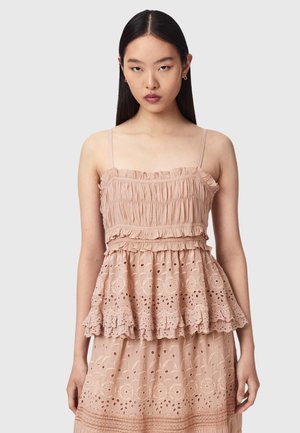 Vrouw met stijl donker haar die een beige mouwloze top draagt met ruches en opengewerkt kantdetail, staand tegen een effen achtergrond.